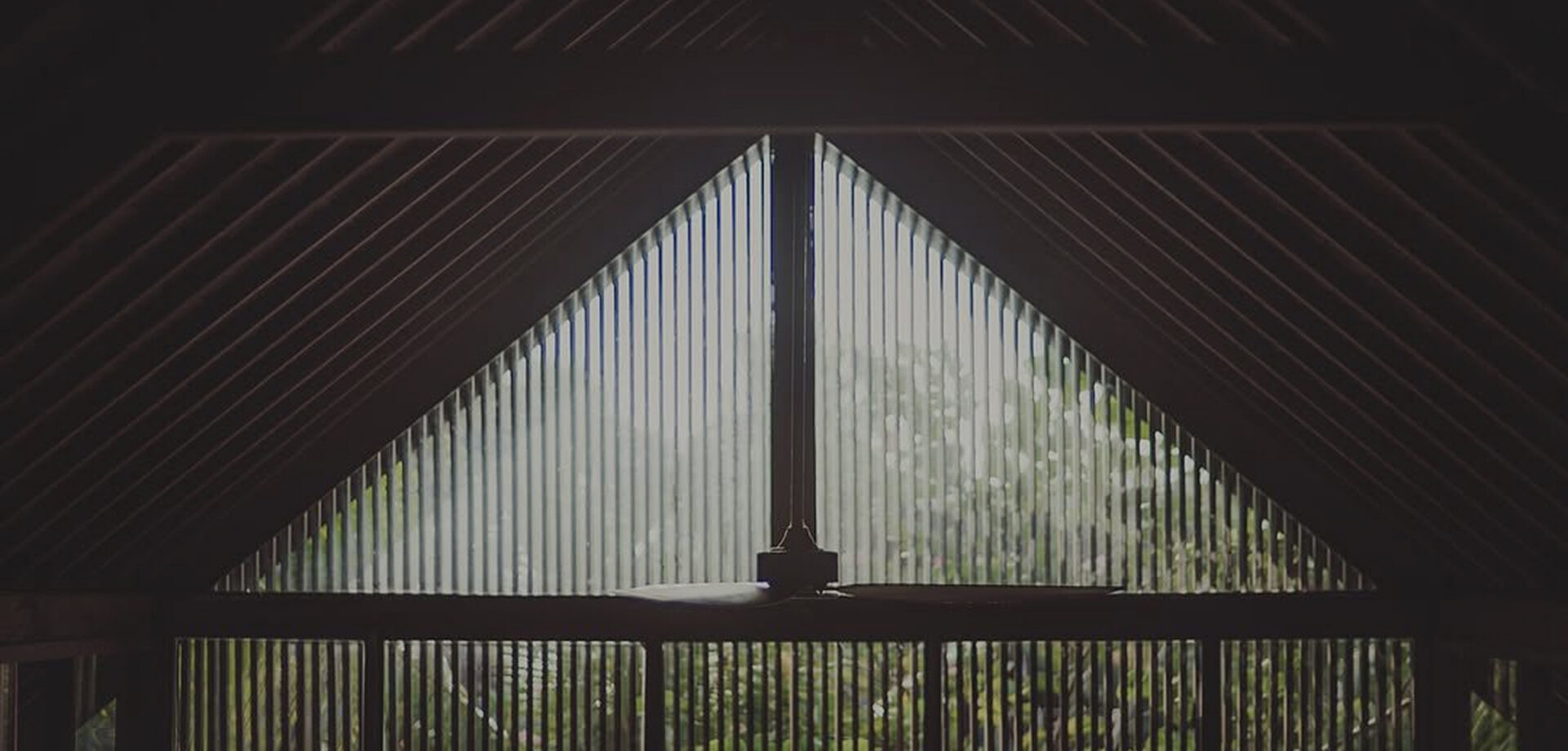 View from inside a dark timber pavilion through triangular gable screens of slats to a green garden