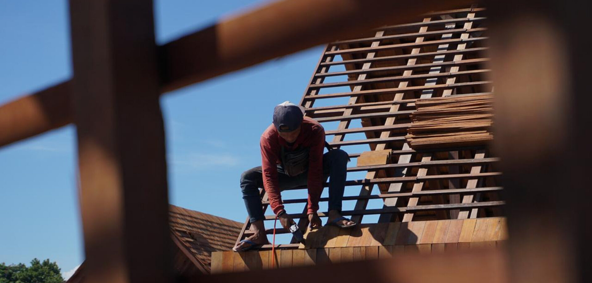 Worker fastening reclaimed hardwood battens on a steep pitched roof frame under a clear blue sky