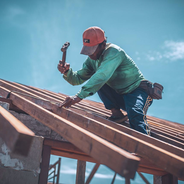 Builder in a green shirt hammering timber battens onto a sloped roof structure under bright sky