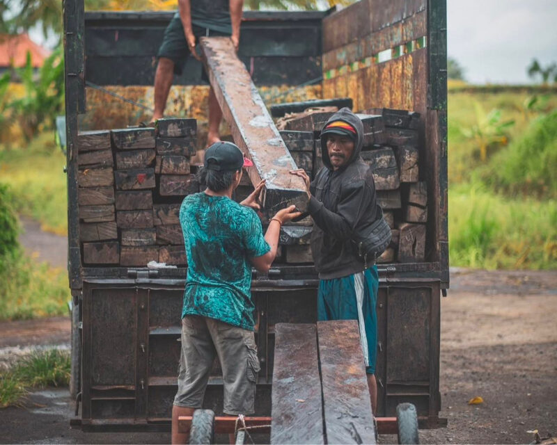 Two workers unloading thick reclaimed ironwood timber blocks from a truck on a rural Bali road