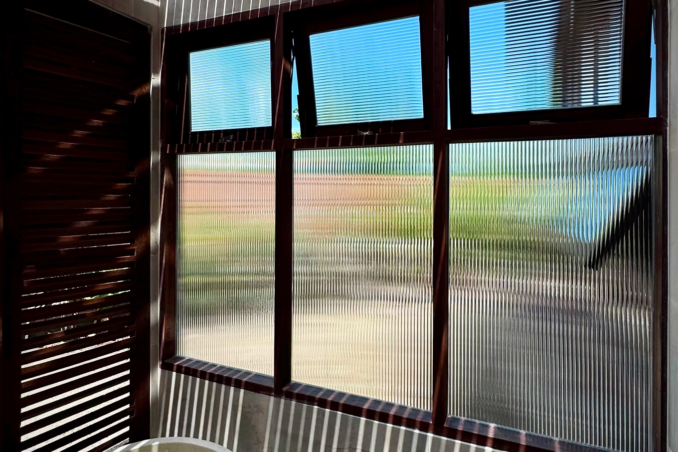 Dark-stained solid wood awning windows with fluted reeded glass framing a green garden view