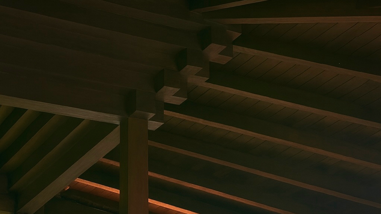 Warm-lit underside of interlocking solid timber roof beams and posts forming an exposed framework