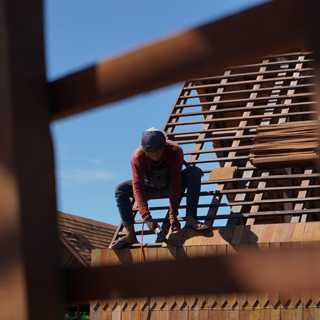 Worker seated on a pitched roof frame fixing reclaimed hardwood battens, framed by timber beams