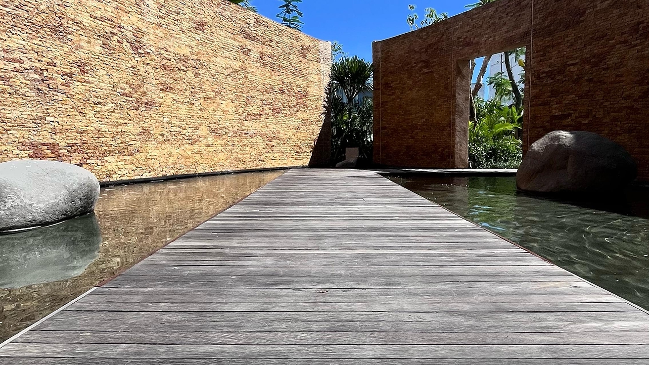 Grey hardwood boardwalk crossing a still reflecting pool toward a sandstone wall and garden