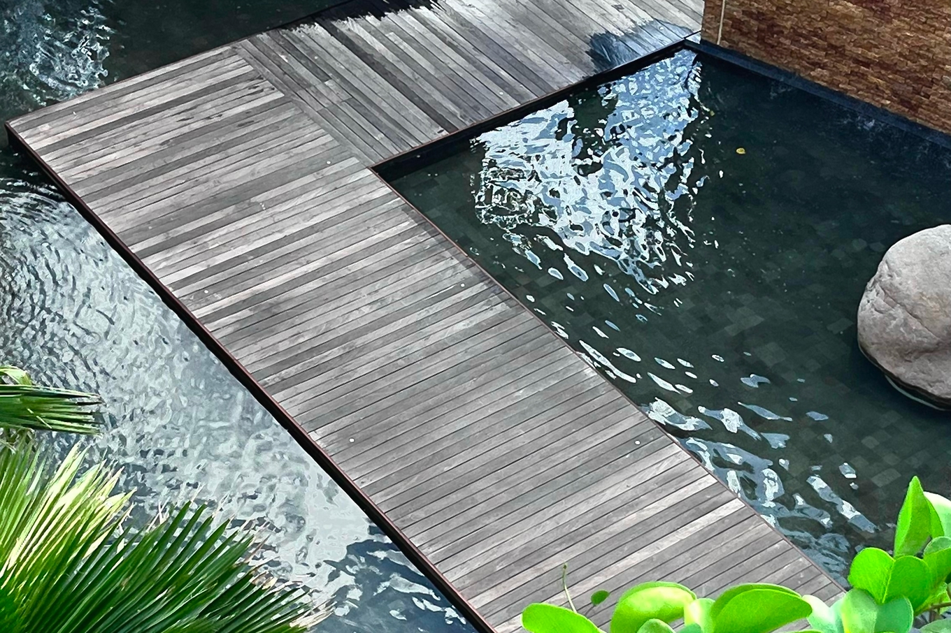 Weathered grey hardwood deck walkway crossing a dark koi pond framed by tropical palms